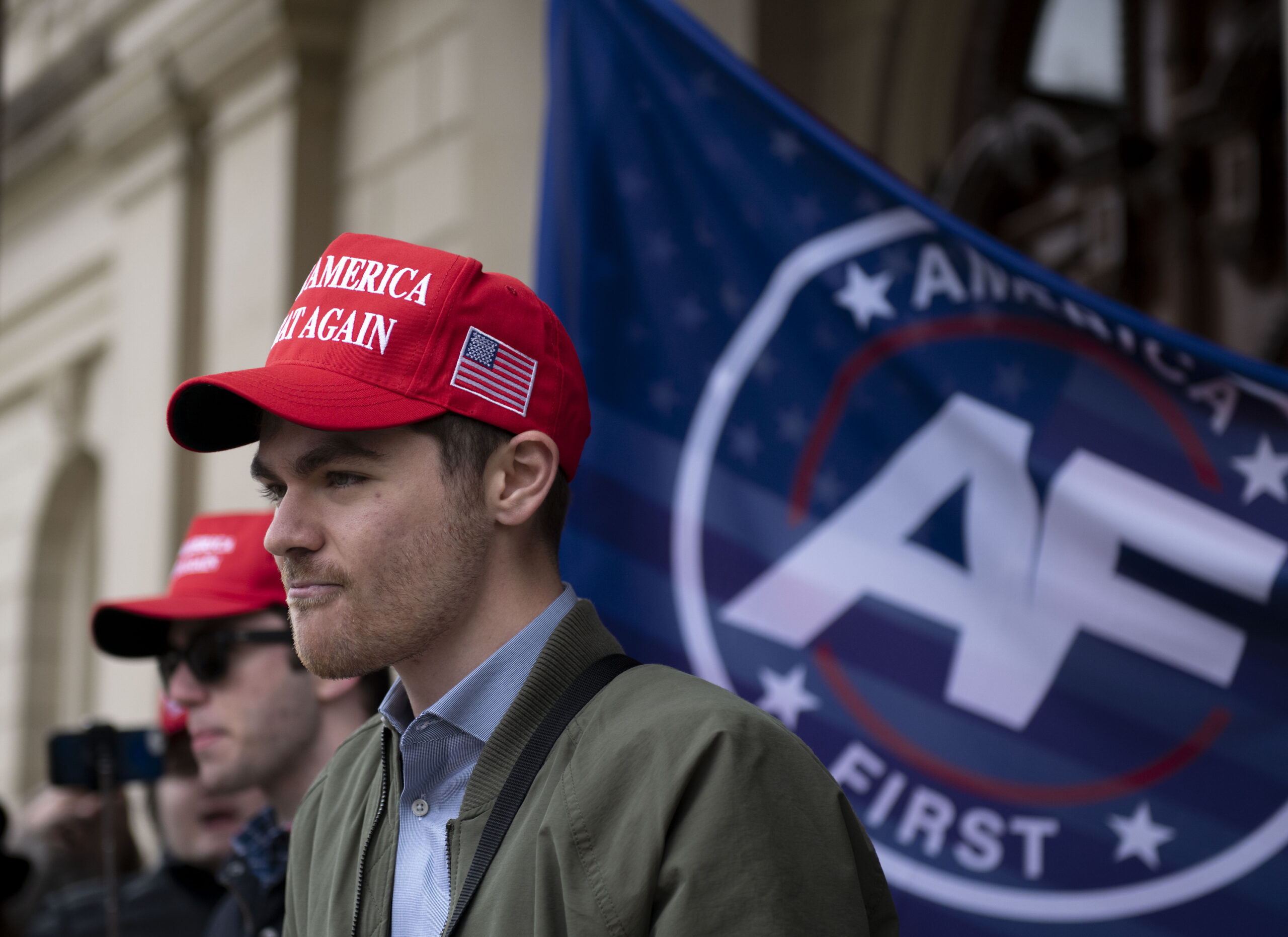 Nick Fuentes wearing a MAGA hat, standing in front of an America First flag.